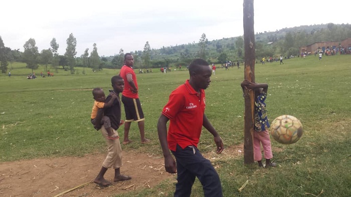 Abantu banyuraga mu kibuga mu gihe abandi barimo gukina. Umwe ahetse umwana mu kibuga, undi yegamye ku izamu